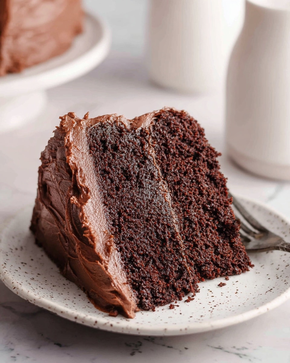 A slice of two-layer chocolate cake sits on a white plate with small black speckles. The cake layers are dark brown and moist with a soft crumb texture. Between the layers is a thick spread of smooth, rich chocolate frosting that also covers the top and side of the slice, with visible swirls and soft peaks. The background shows a blurred white marbled texture surface and a couple of white containers. photo taken with an iphone --ar 4:5 --v 7