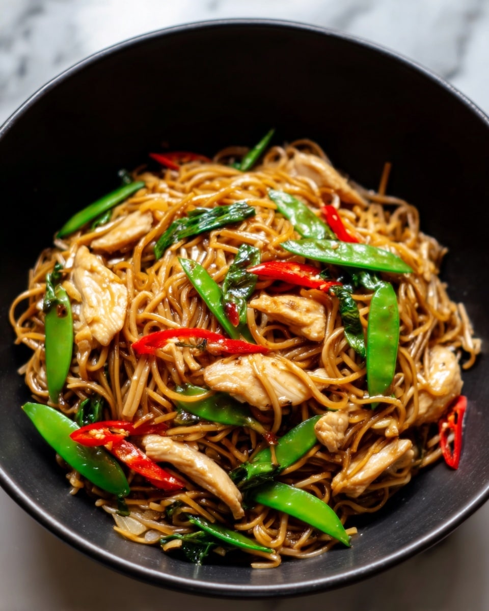 A black bowl filled with stir-fried noodles mixed with pieces of cooked chicken, bright green snap peas, and thin red chili slices. The noodles have a shiny texture, coated in a brown sauce, with the vegetables and chicken evenly spread through the dish. The background shows a white marbled surface. Photo taken with an iphone --ar 4:5 --v 7