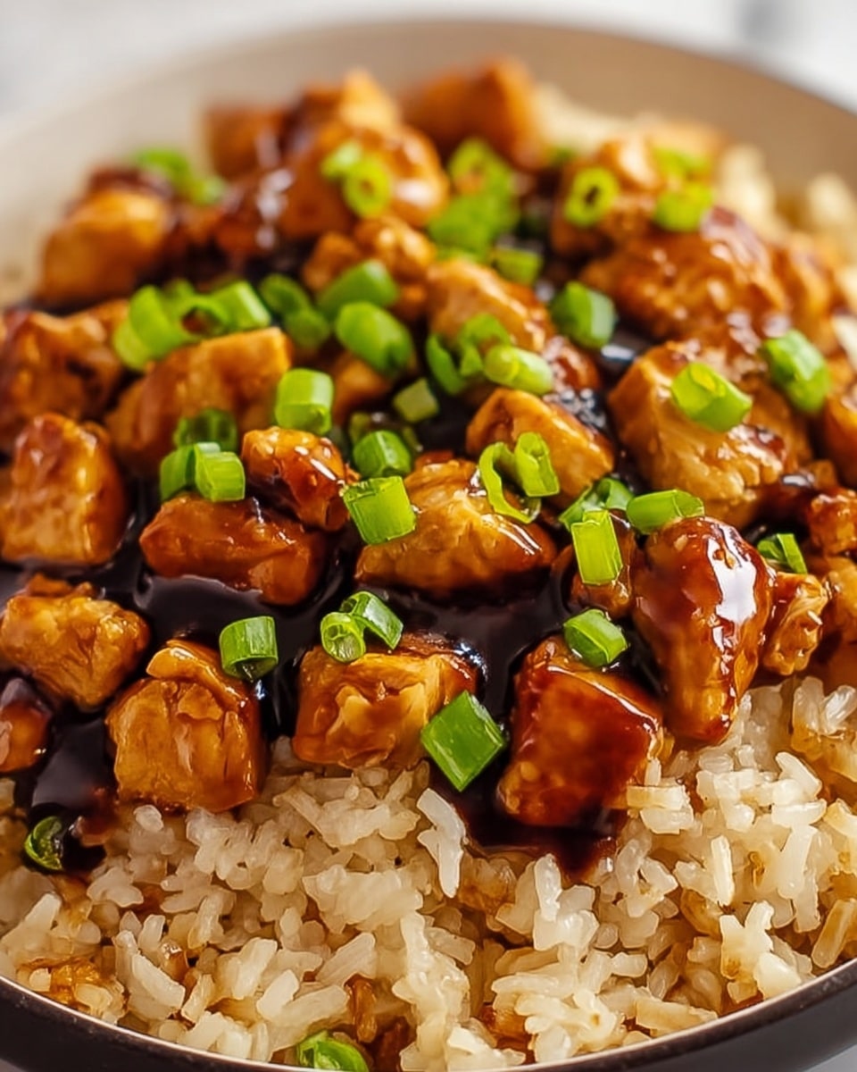 A close-up view of a dish with two main layers, served in a white bowl on a white marbled surface. The bottom layer is light brown fried rice with a slightly sticky texture, showing individual grains mixed with small bits of vegetables. On top, cubed chicken pieces are coated in a shiny dark brown sauce that looks thick and glossy. Bright green chopped scallions are sprinkled evenly over the chicken, adding a fresh contrast to the caramel color of the sauce. The photo taken with an iphone --ar 4:5 --v 7