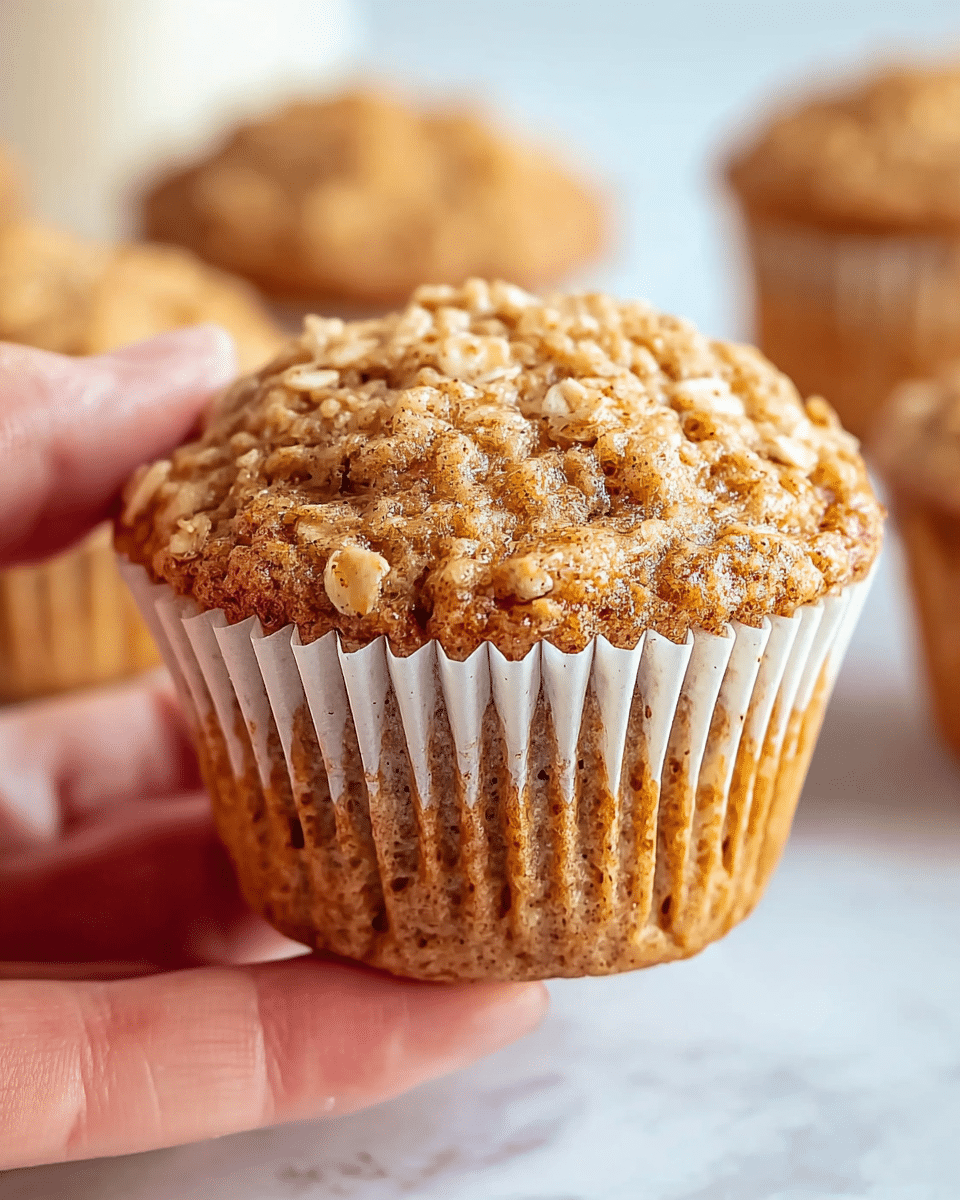 A close-up view of a single oatmeal muffin held between a woman's thumb and forefinger, showing a golden-brown textured top with visible whole oats sprinkled throughout, creating a slightly rough and crumbly surface. The muffin liner is white with vertical ridges, hugging the base snugly and slightly translucent to reveal the muffin's warm, moist interior. In the softly blurred background, there are more muffins placed on a white marbled surface, adding depth to the image. The lighting highlights the muffin's crisp edges and oats, giving it a fresh, homemade look. photo taken with an iphone --ar 4:5 --v 7