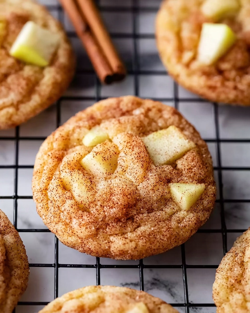 The image shows several round cookies on a black cooling rack placed over a white marbled surface. Each cookie has a golden-brown base with a slightly puffy texture and is sprinkled with fine cinnamon and sugar on top. Chunks of pale apple pieces are embedded in the surface of the cookies. One cookie on the top right also has a cinnamon stick resting on it, adding a rustic touch. The cookies look soft and chewy with a gentle crumbly edge. Photo taken with an iphone --ar 4:5 --v 7