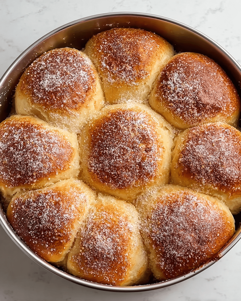 A round baking pan filled with nine soft, golden-brown rolls that are puffy and touching each other closely. Each roll has a slightly shiny surface with a dusting of fine white powdered sugar on top, creating a light, snowy effect. The rolls show a slight gradient of color, darker and crispier on top while lighter and fluffier on the sides. The pan edges show slight baking marks and the texture of the rolls looks airy and tender. The baking pan is set against a white marbled texture background. photo taken with an iphone --ar 4:5 --v 7