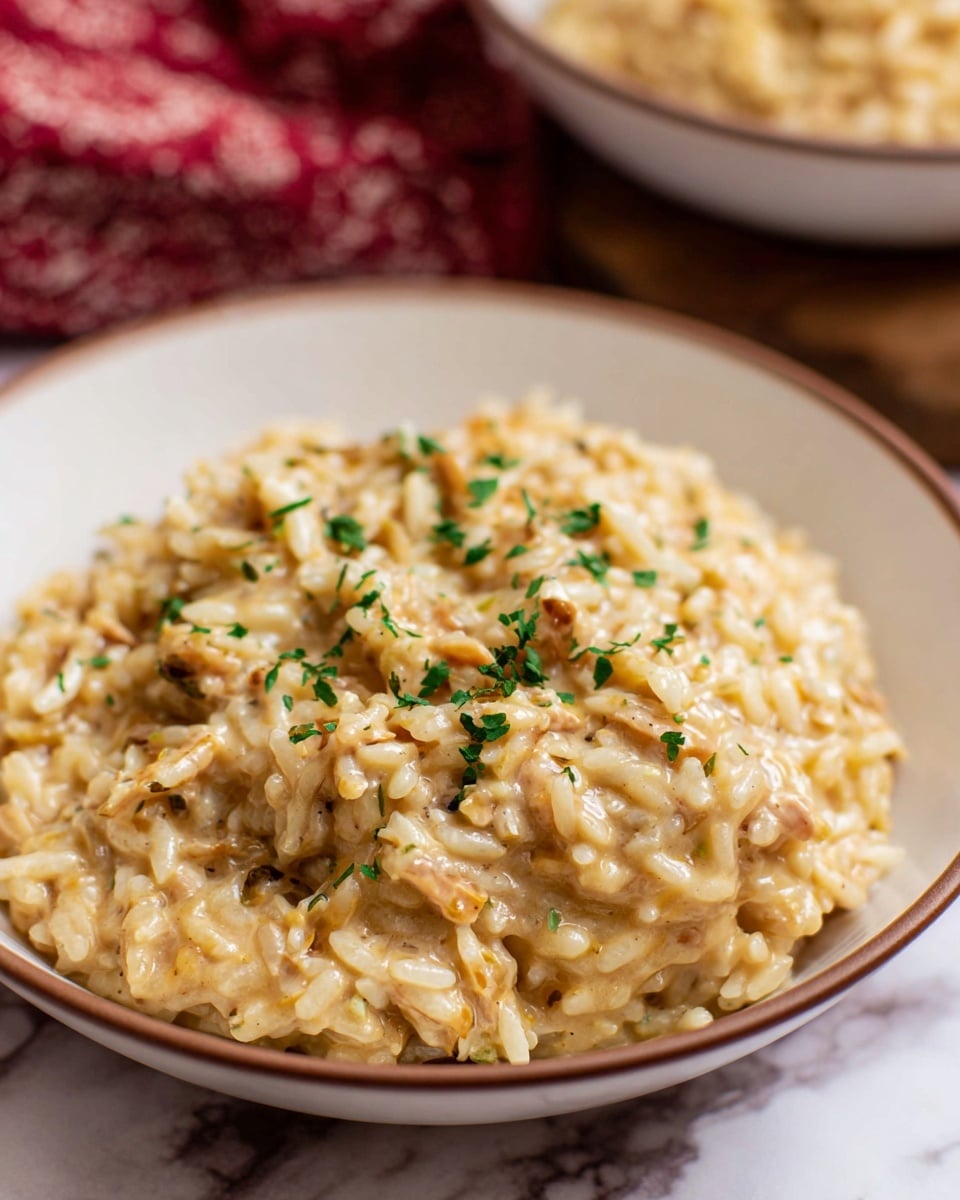 A close-up view of a creamy rice dish served in a white bowl with a brown rim, showing soft, mixed rice grains coated in a rich, light golden sauce that looks smooth and slightly thick. The dish is garnished with small pieces of green herbs scattered on top, adding a touch of color. The background is a blurred white marbled texture with hints of other bowls and a red patterned cloth. photo taken with an iphone --ar 4:5 --v 7