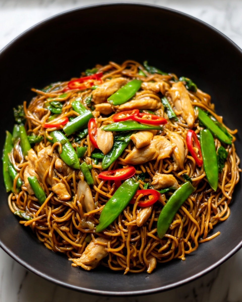 A black bowl filled with stir-fried noodles, showing three main layers: the bottom layer has thin, brown noodles tangled together; on top of the noodles are pieces of light golden-brown chicken scattered around; the top layer includes bright green snap peas and thin slices of red chili peppers spread evenly. The noodles look shiny with sauce, and the vegetables add a fresh pop of color. The bowl is set on a white marbled surface. photo taken with an iphone --ar 4:5 --v 7