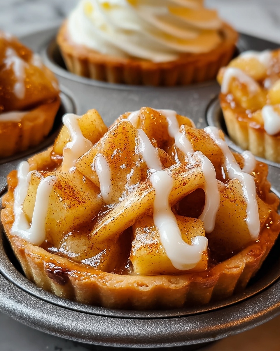 The image shows a close-up of a small tart in a metal muffin tray. The tart has one layer of golden-brown crust forming its base and edge. On top of the crust are irregular pieces of cooked apple, colored light brown with a shiny glaze, suggesting they are coated with cinnamon and sugar. The apples are sprinkled lightly with cinnamon powder. Over the apples, there are smooth, thick white icing lines drizzled in a zig-zag pattern. In the background, there is another similar tart with a swirl of white cream on top, hinting at a soft texture. The overall look is warm and inviting, with the tray resting on a white marbled surface. photo taken with an iphone --ar 4:5 --v 7