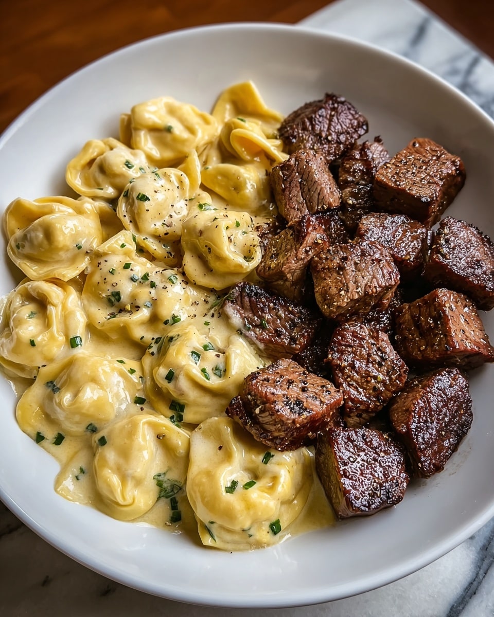 The image shows a white plate filled with two main layers. On the left side, there is a creamy layer of cheese-filled tortellini pasta, covered in a light yellow sauce with a smooth, slightly glossy texture and sprinkled with small green herb pieces and black pepper. On the right side, there is a layer of evenly cut, browned steak cubes, with a crispy, seared outer texture in dark brown shades with some seasoning visible. The plate is set on a white marbled surface with wood partially visible on one corner. photo taken with an iphone --ar 4:5 --v 7