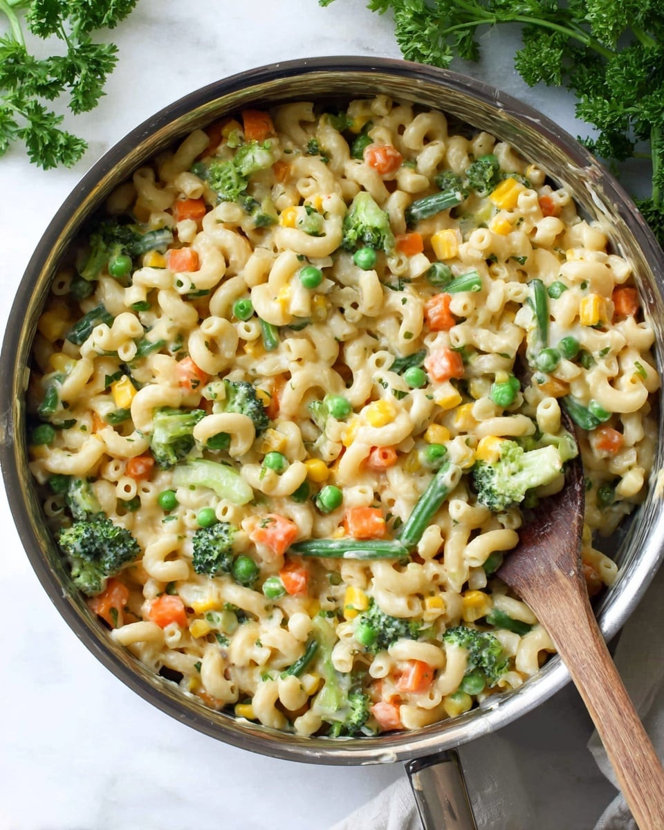 A close-up of a shiny metal pan filled with creamy macaroni pasta mixed with bright green broccoli florets, peas, and other small diced vegetables like orange carrots and yellow corn, all coated in a smooth, light-colored sauce. A wooden spoon rests on the right side inside the pan, and fresh green herb leaves are visible at the top left corner on a white marbled surface underneath. photo taken with an iphone --ar 4:5 --v 7