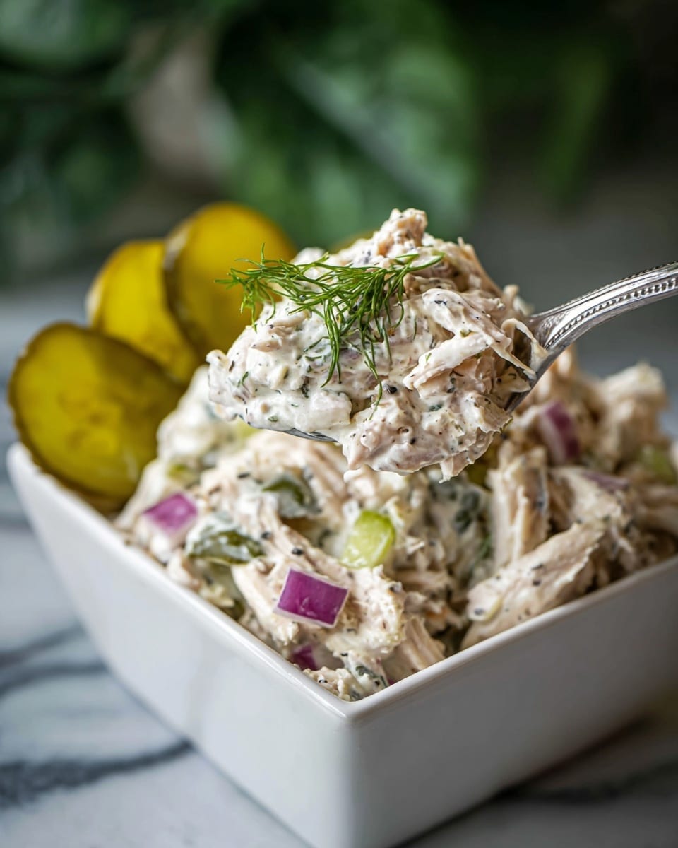 A close-up image of a spoonful of creamy chicken salad held over a white square bowl filled with the same chicken salad. The chicken salad has three main visible layers: shredded white chicken meat mixed with a creamy, speckled dressing, small chunks of purple-red onion, and small pieces of green celery and fresh dill. In the background inside the bowl, there are several thin slices of yellow-green pickles arranged at the back. The surface under the bowl is a white marbled texture. The photo taken with an iphone --ar 4:5 --v 7