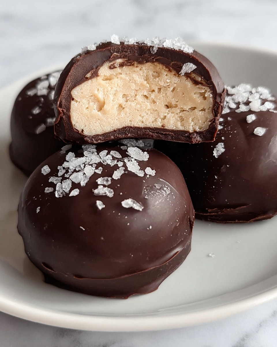 The image shows three chocolate-covered truffles on a white plate with a white marbled texture background. Two truffles are whole, round, and have a smooth dark brown chocolate coating topped with coarse sea salt flakes for added texture and shine. The third truffle is cut in half and placed on top of the two whole truffles, revealing a thick outer dark chocolate shell and a creamy, light tan filling inside with a slightly grainy texture. The contrast between the dark chocolate outer layer and the pale filling is clear, with the sea salt flakes scattered on top of the chocolate shells. photo taken with an iphone --ar 4:5 --v 7