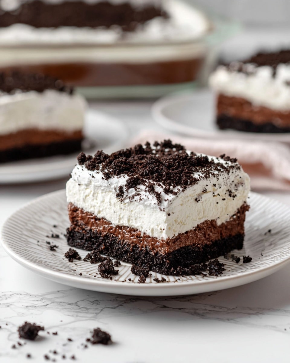 A square piece of layered dessert sits on a white plate with a patterned rim, placed on a white marbled surface. The dessert has four visible layers: a dark, crumbly chocolate cookie crust at the bottom, followed by a thick white creamy layer, then a smooth dark chocolate layer, topped with a fluffy white whipped cream layer, and finished with a generous layer of crushed dark chocolate cookie crumbs. Some crumbs are scattered around the dessert on the plate. In the background, more pieces of the dessert are visible on white plates and in a glass dish. Photo taken with an iphone --ar 4:5 --v 7
