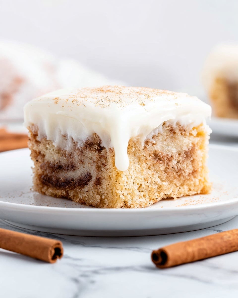 A single square piece of cinnamon roll cake is placed on a white plate. The cake has two visible layers: the bottom layer is light brown with swirls of darker cinnamon spice creating a textured and slightly crumbly look, while the top layer is a smooth, thick white frosting that drips slightly down the sides. The background is a white marbled texture, and there are two cinnamon sticks placed in front of the plate, slightly out of focus. Photo taken with an iphone --ar 4:5 --v 7